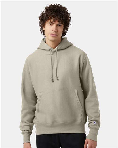 A young person with curly hair wears a light gray Champion S101 Reverse Weave hoodie with a front pocket, standing against a plain white background.