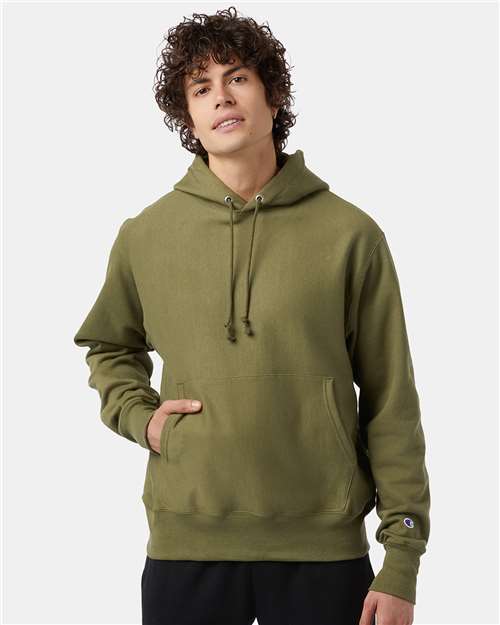 A person with curly brown hair wears an olive green Champion S101 hoodie with a front pocket, standing against a plain light background—one hand in the pocket, the other at their side.