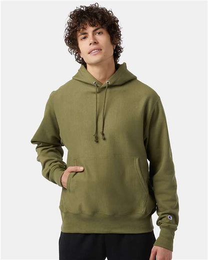 A person with curly brown hair wears an olive green Champion S101 hoodie with a front pocket, standing against a plain light background—one hand in the pocket, the other at their side.