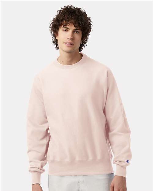 A young man with curly dark hair wears a light pink Champion S149 sweatshirt and light pants, standing against a plain white background.