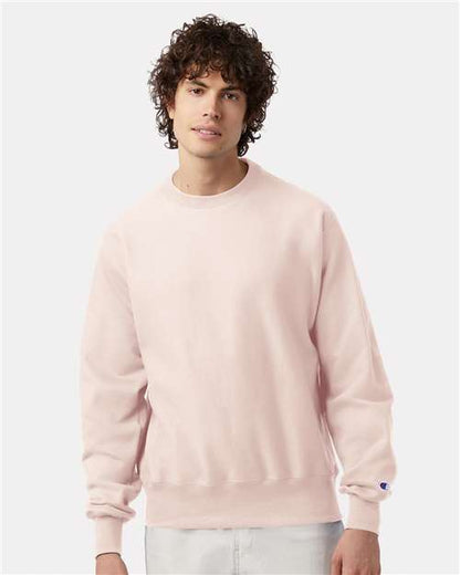 A young man with curly dark hair wears a light pink Champion S149 sweatshirt and light pants, standing against a plain white background.