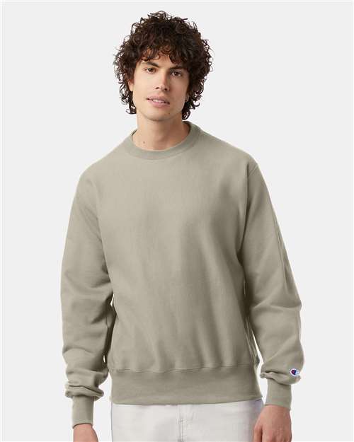 A young man with curly brown hair wears a light gray Champion S149 sweatshirt and white pants, standing against a plain light background.