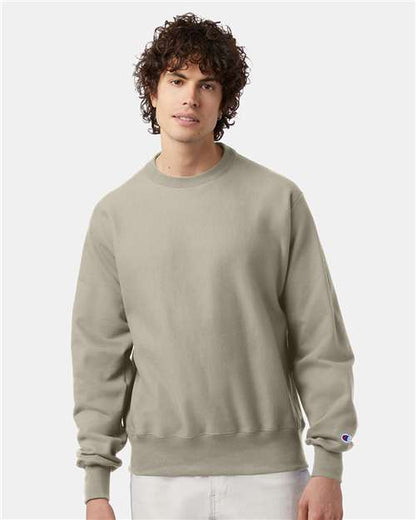 A young man with curly brown hair wears a light gray Champion S149 sweatshirt and white pants, standing against a plain light background.