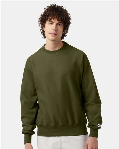 A young man with curly hair is wearing a Champion S149 olive green sweatshirt and light-colored pants, standing against a plain light gray background.