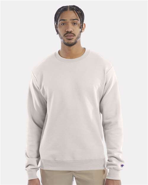 A man with short braided hair and a mustache wears a light beige Champion S600 sustainable sweatshirt made from recycled polyester and tan pants, standing against a plain light gray background.