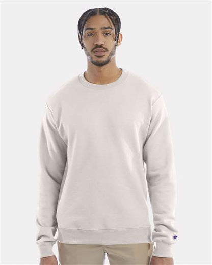 A man with short braided hair and a mustache wears a light beige Champion S600 sustainable sweatshirt made from recycled polyester and tan pants, standing against a plain light gray background.