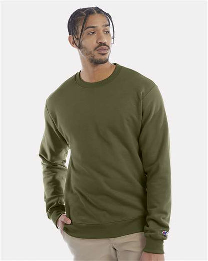A man with braided hair wears a Champion S600 sustainable sweatshirt in olive green and beige pants, standing with one hand in his pocket and looking slightly to the side against a plain light background.
