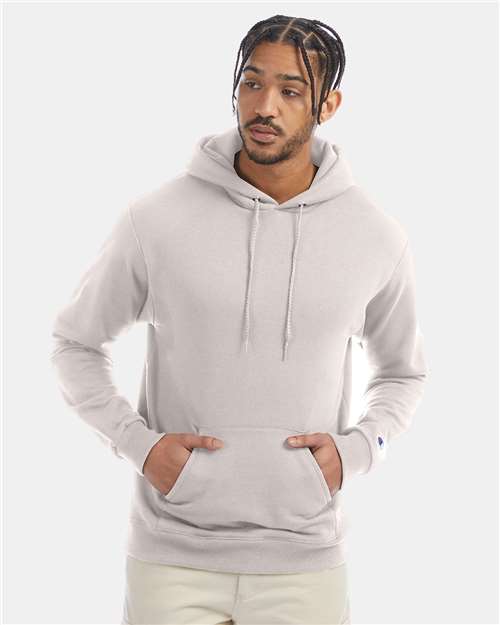 A man with braided hair wears a light gray Champion S700 hoodie, made from recycled polyester, hands in the front pocket, standing against a plain light background.