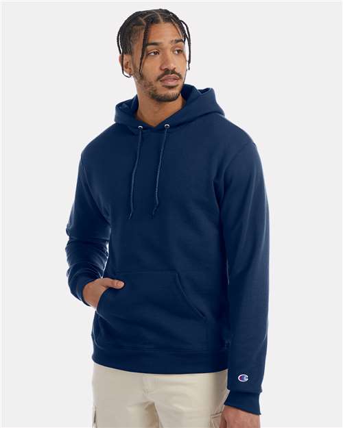 A man with short braids wears a navy blue Champion S700 hoodie made from recycled polyester, featuring a front pocket and sleeve logo, standing against a plain light background and looking slightly to the side.