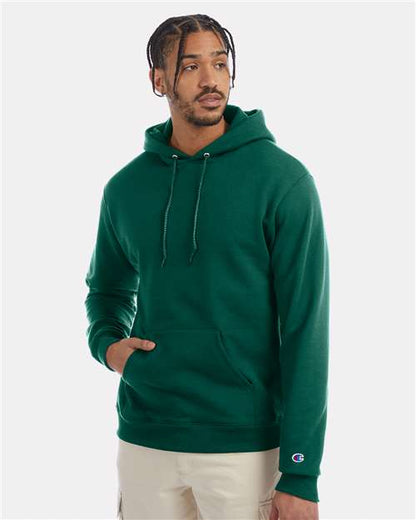 A man wears a green Champion S700 hoodie made from recycled polyester, featuring drawstrings and a front pocket, as he stands against a plain white background looking slightly to his right.