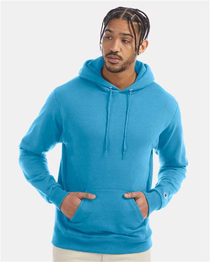 A man with braided hair wears the Champion S700 hoodie in bright blue, made from recycled polyester, standing against a plain light background and gazing slightly to the side with a neutral expression.