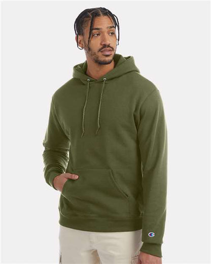 A man wears a green Champion S700 hoodie made from recycled polyester with a front pocket and beige pants, standing against a plain light background and looking to his left.