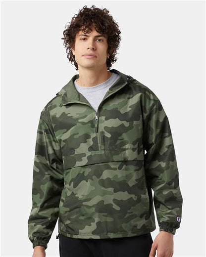 A young man with curly hair wears the Champion CO200 wind- and water-resistant jacket in green camo over a gray shirt, standing against a plain light background.