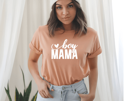 A woman indoors, in soft light with white curtains and a plant, wears the peach-colored Boy mama T-shirt by DB Custom Creations, featuring boy MAMA and a heart design created with 11-inch DTF printing.