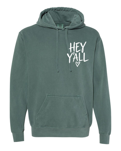 Blue Spruce Hey Ya’ll Comfort Color Hooded Sweatshirt featuring a front kangaroo pocket and the text HEY YALL with a small heart icon printed on the left chest.