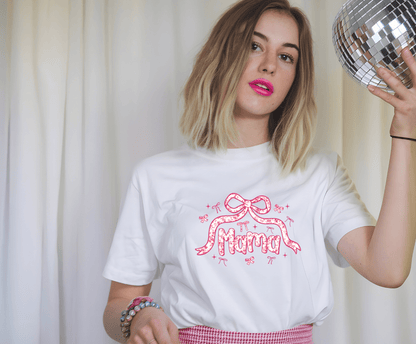 A young woman with shoulder-length blonde hair wears the Coquette Mama white T-shirt by DB Custom Creations, featuring a vibrant Mama and pink bow graphic. She pairs it with a pink skirt and colorful bracelets, holding a disco ball.