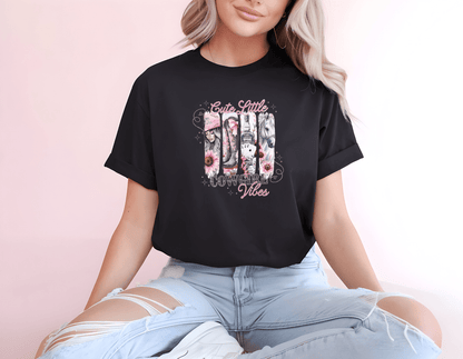 A woman with long blonde hair wears a black DB Custom Creations Cute Cowgirl Vibes DTF-printed t-shirt with pink floral designs, sitting cross-legged in ripped light blue jeans against a pale pink background.