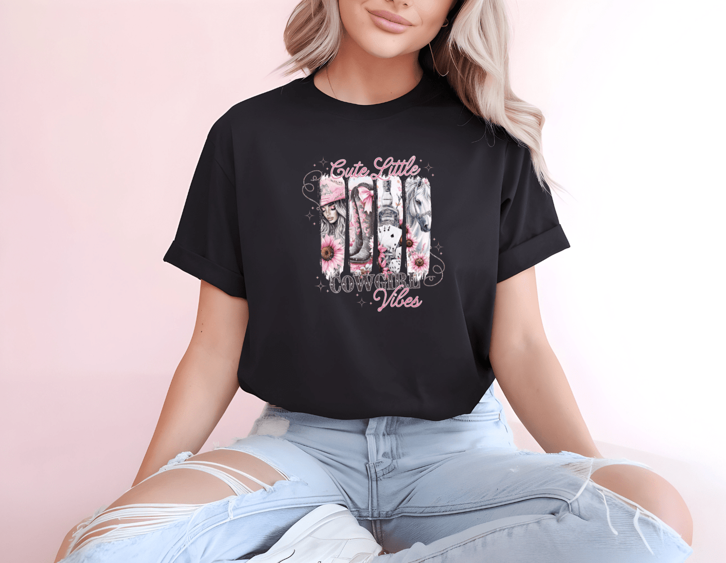 A woman with long blonde hair wears a black DB Custom Creations Cute Cowgirl Vibes DTF-printed t-shirt with pink floral designs, sitting cross-legged in ripped light blue jeans against a pale pink background.