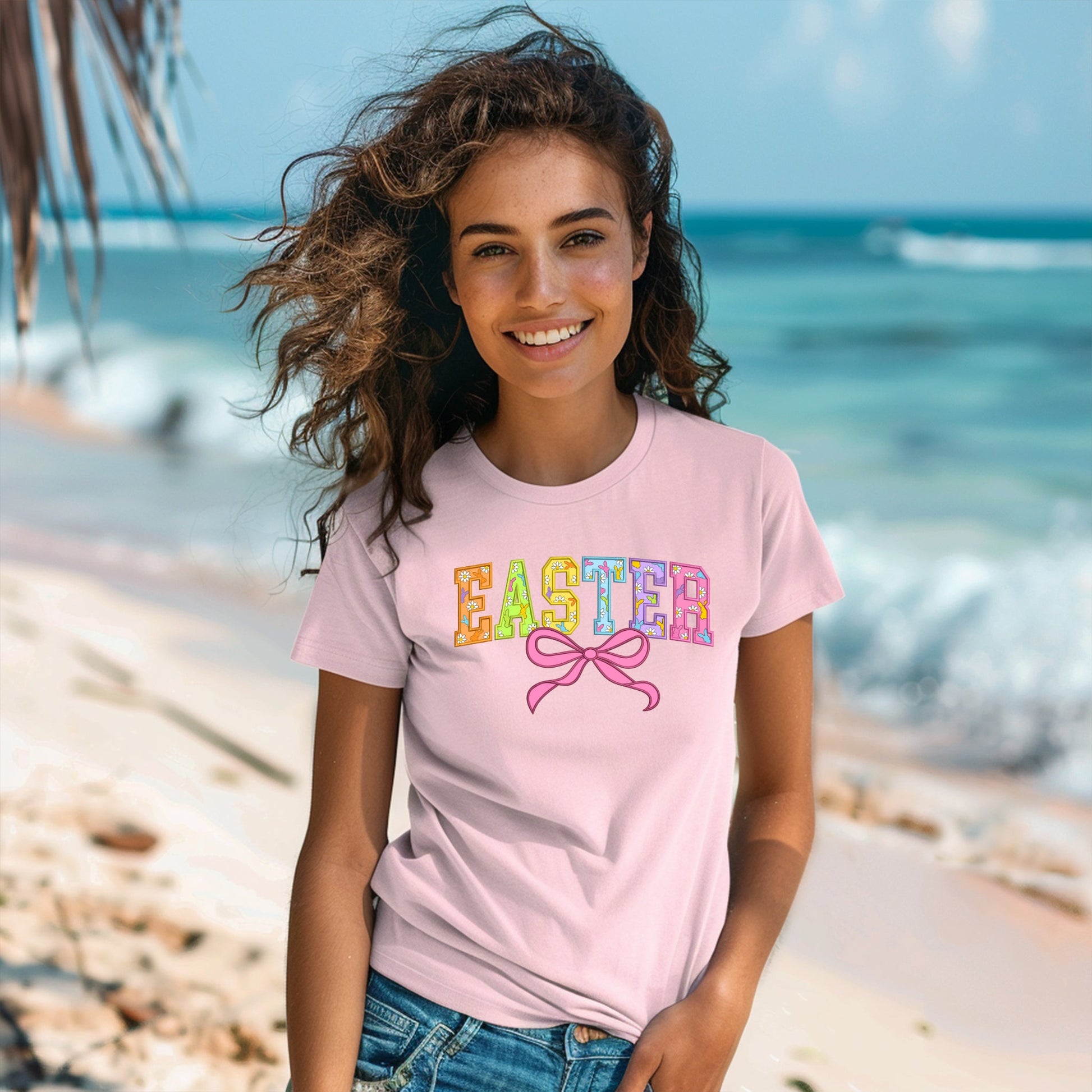 A smiling woman with curly hair stands on a sunny beach wearing DB Custom Creations Easter light pink T-shirt, featuring vibrant Easter letters and a pink bow in DTF print, with the ocean and sky in the background.