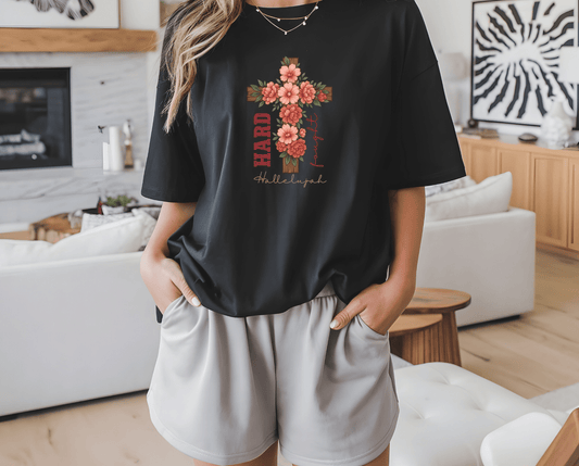 A woman stands in a modern living room, wearing DB Custom Creations Floral Cross Hard Fought Hallelujah black T-shirt with 11-inch DTF floral cross graphic and text, paired with light shorts and her hands in her pockets.