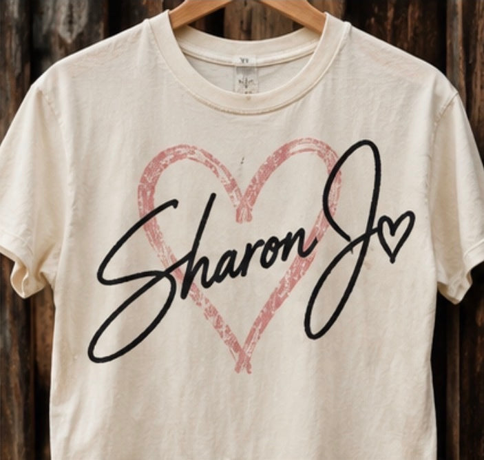 The Sharon Jo Signature T-shirt by DB Custom Creations is a cream-colored Comfort Colors 1717 tee displayed on a hanger, featuring a large hand-drawn pink heart outline and the words “Sharon Jo” in bold black script across the front. Made from soft ring-spun cotton and garment dyed for a vintage look, it offers both comfort and timeless style.