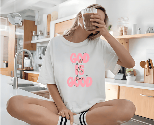 A woman sits cross-legged on a bright kitchen counter, holding a coffee cup. She wears the “God is good Pink” T-shirt by DB Custom Creations, featuring vibrant DTF printing in pink and black text.