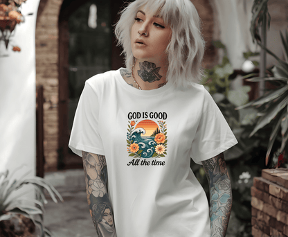 A gray-haired, tattooed woman wears a white T-shirt from DB Custom Creations featuring the God is good DTF Transfer design with waves, flowers, and a sunset. She stands outside near plants and brick walls.