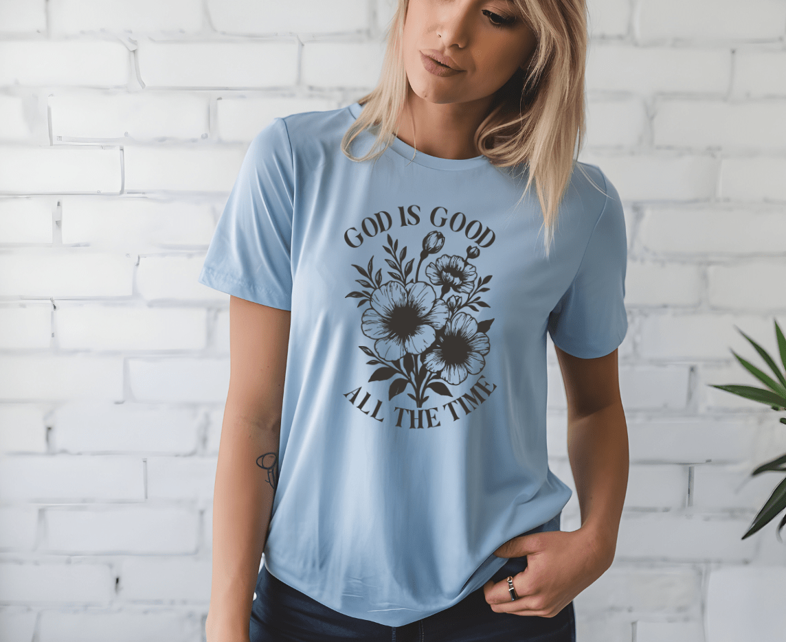 A woman models the “God is good” t-shirt by DB Custom Creations, featuring a black floral design and sleek 11-inch DTF printing, standing in front of a white brick wall with one hand in her pocket.
