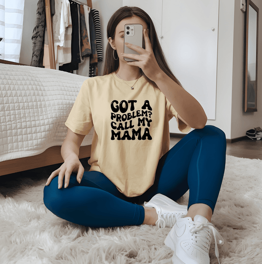 A young woman takes a mirror selfie on her bedroom floor wearing the Got a problem call my mama t-shirt by DB Custom Creations, paired with blue leggings and white sneakers; a bed and clothes rack are visible in the background.