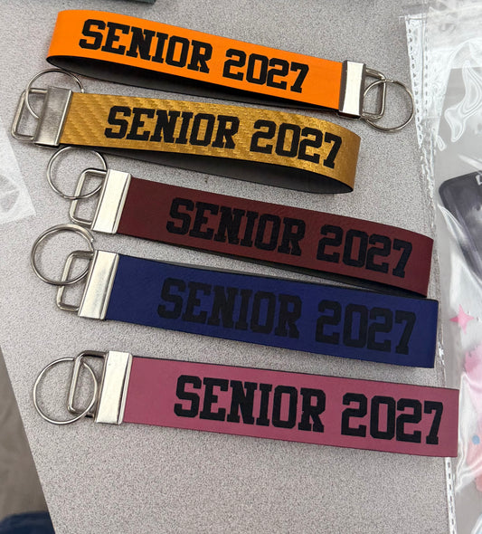 Five Custom Laser Engraved Key Chains from DB Custom Creations, each in a different color—orange, gold, maroon, navy blue, and pink—are displayed on a gray surface. Featuring “Senior 2027” in black or gold lettering, these personalized keychains make ideal graduation gifts to celebrate the class of 2027.