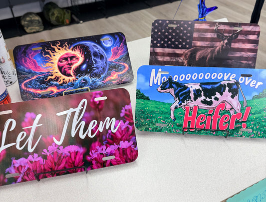 Here’s a revised version using your product data: Four Custom UV Printed License Plates by DB Custom Creations on display: one features psychedelic sun and moon artwork, another displays a bull with an American flag, a third reads “Let Them” over pink flowers, and the last showcases a cow with the phrase “Mooove over Heifer!”—each adding a personalized touch to your vehicle.