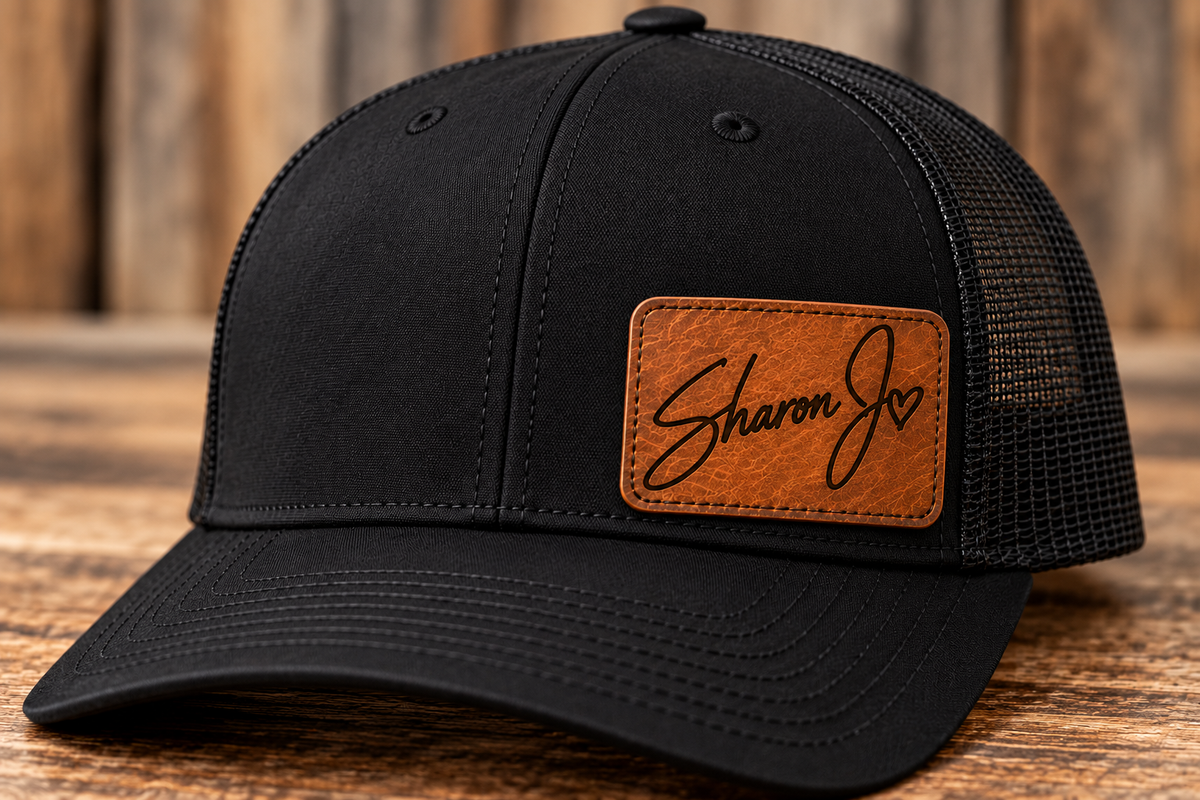 The Sharon Jo Signature Hat by DB Custom Creations, a stylish black mesh-back Richardson 112 baseball cap with a brown leather patch on the front featuring the handwritten text “Sharon Jo” and a small heart, rests on a wooden surface. This cap also offers an adjustable fit with its convenient snapback closure.
