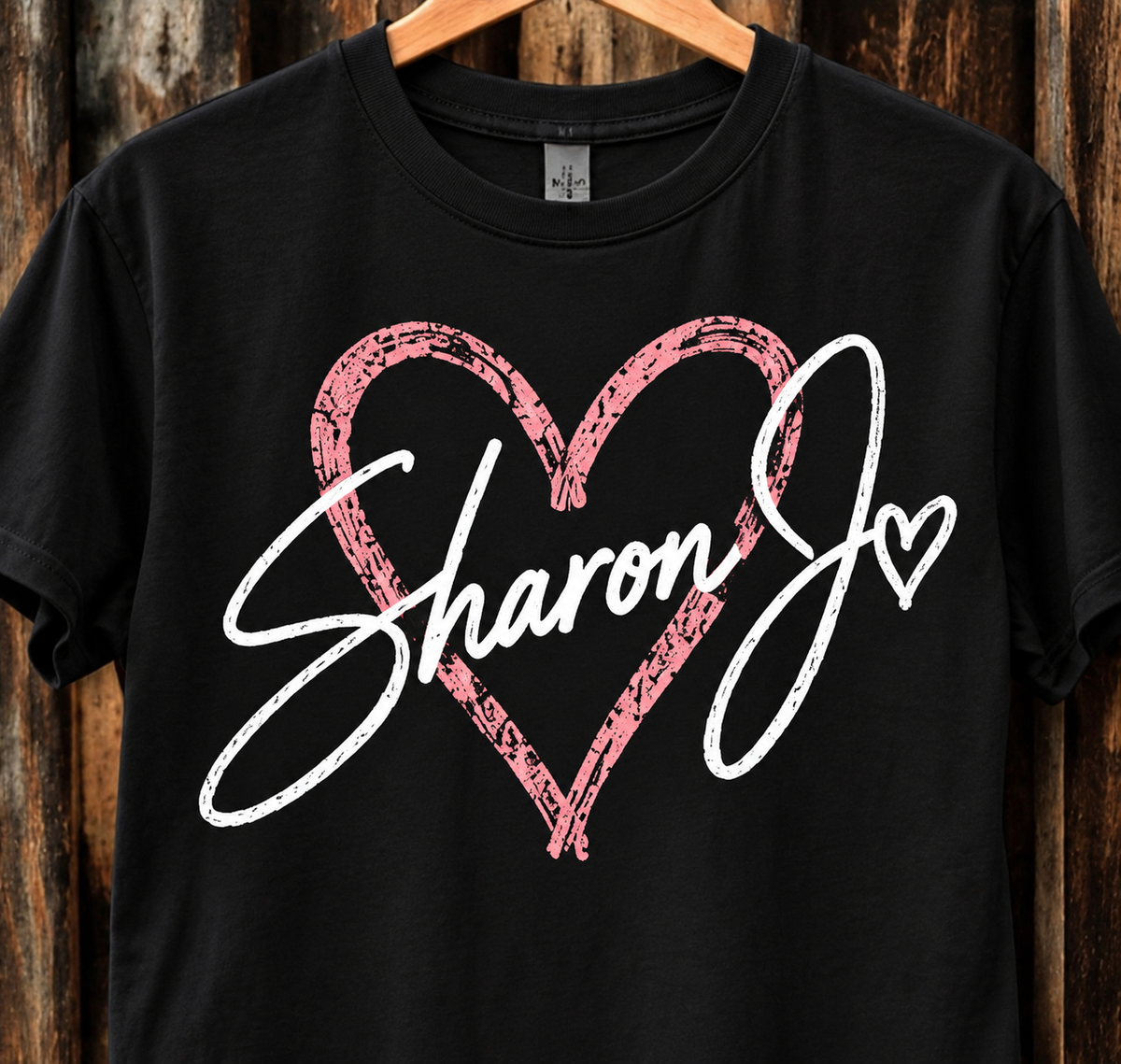 The DB Custom Creations Sharon Jo Signature t-shirt features “Sharon J” in white script across a large, pink, distressed heart outline, with a small heart accenting the letter J. Made from soft ring-spun cotton for a premium feel, this Comfort Colors 1717 garment-dyed tee is displayed on a wooden hanger against a rustic wood background.