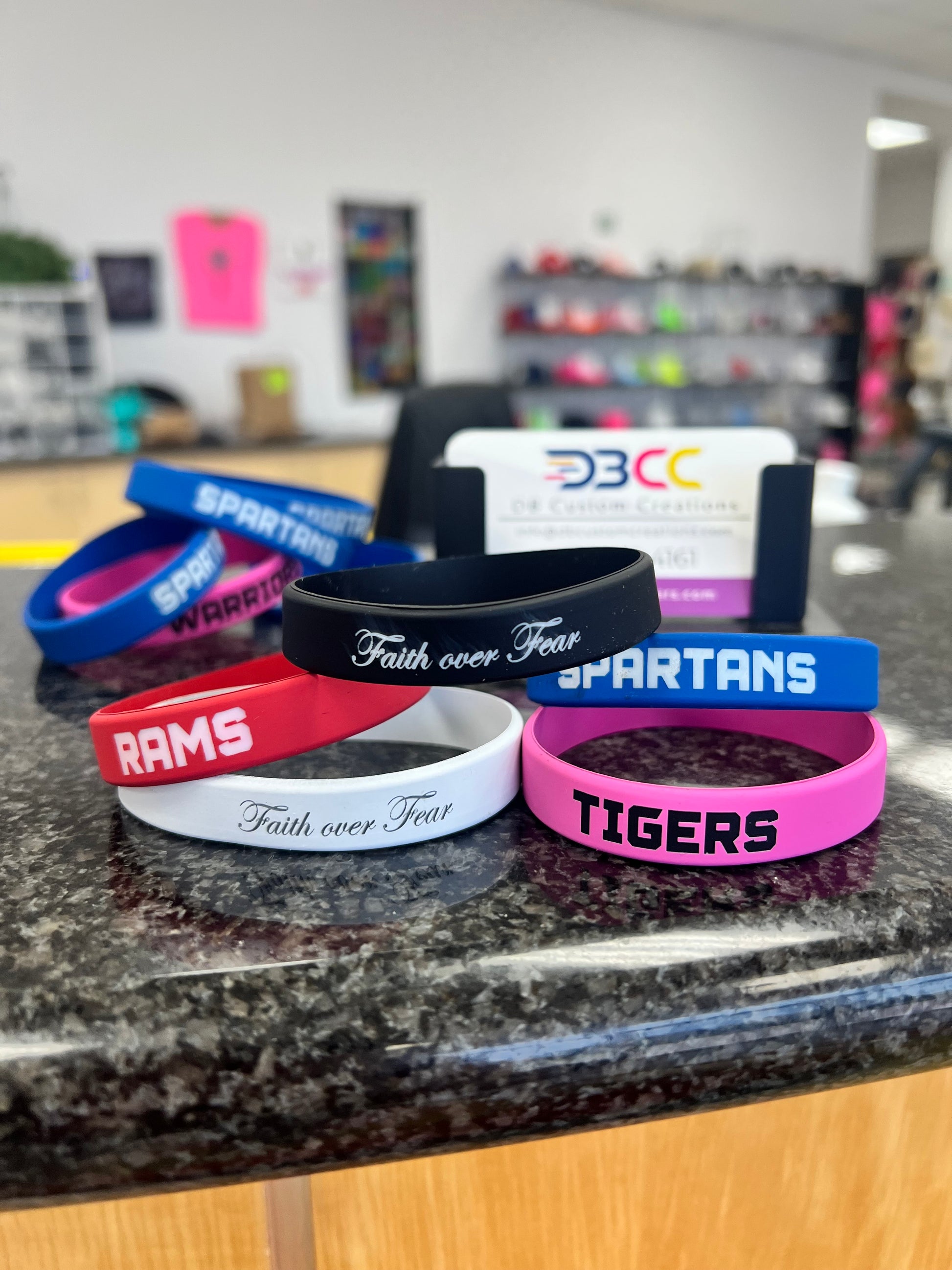 Custom Silicone Bracelets by DB Custom Creations—engraved with team names like RAMS, SPARTANS, TIGERS and quotes such as Faith over Fear—are stacked on a countertop, making a great fundraiser idea. More bands and a card holder appear blurred in the background.