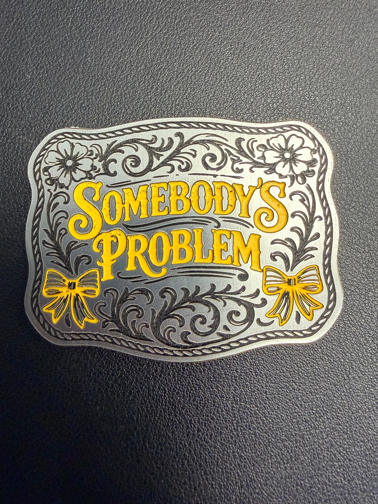 A tri-layered acrylic patch by DB Custom Creations features ornate silver and yellow floral designs and “Somebodys Problem” in bold yellow, accented with bows—ideal for hats using a heat press or adhesive. The plaque lies on a textured dark background.