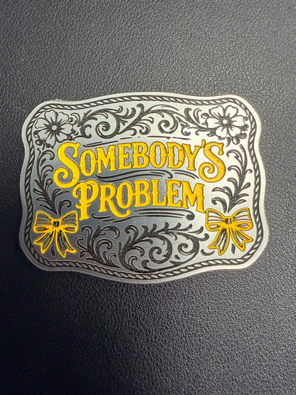 A tri-layered acrylic patch by DB Custom Creations features ornate silver and yellow floral designs and “Somebodys Problem” in bold yellow, accented with bows—ideal for hats using a heat press or adhesive. The plaque lies on a textured dark background.