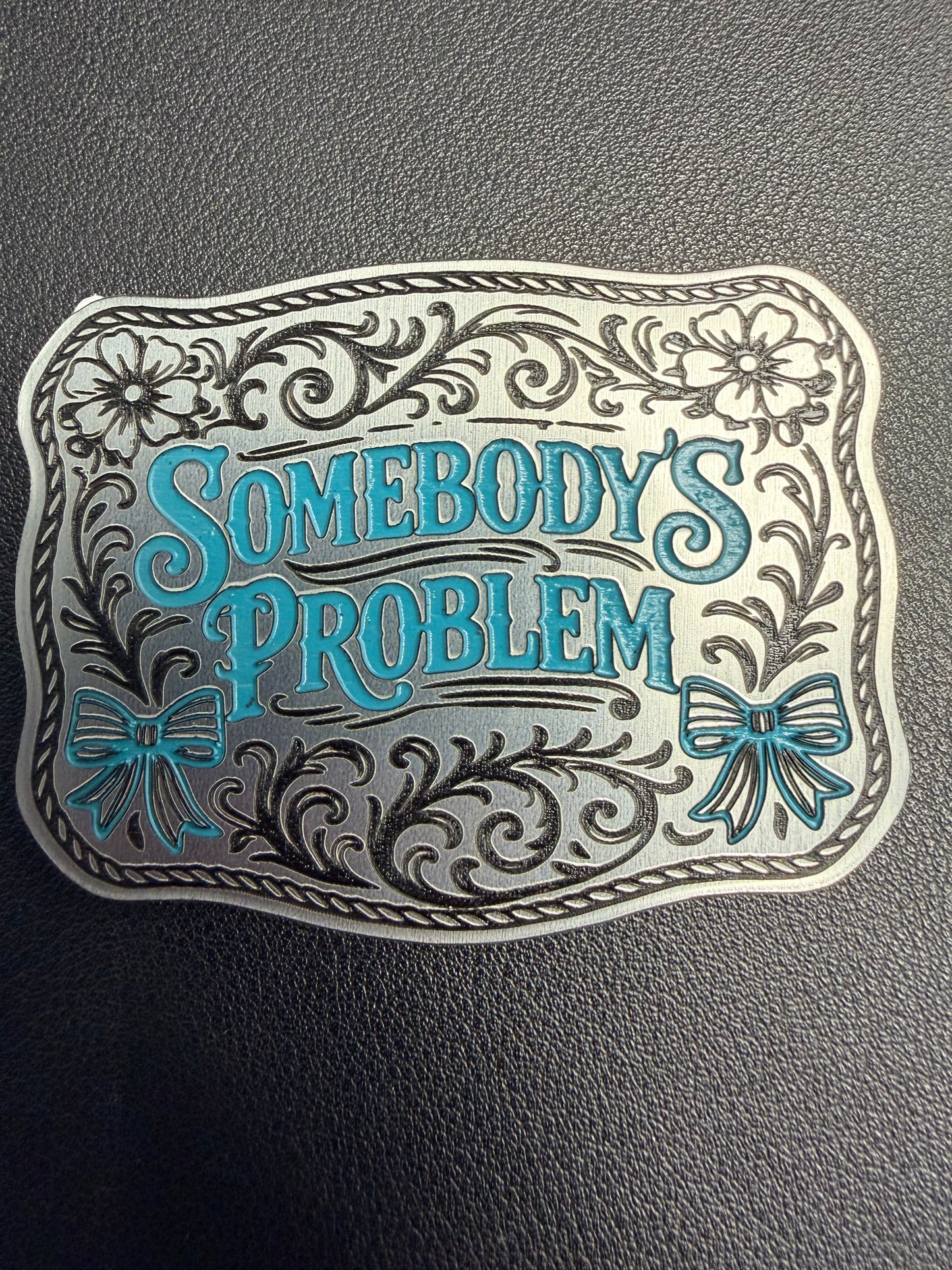 The Custom Laser Engraved Tri Layered Acrylic Patches for Hats by DB Custom Creations feature a decorative, floral design with blue bows and the phrase "Somebodys Problem"—a perfect accessory for fans of unique, high-quality hat patches.