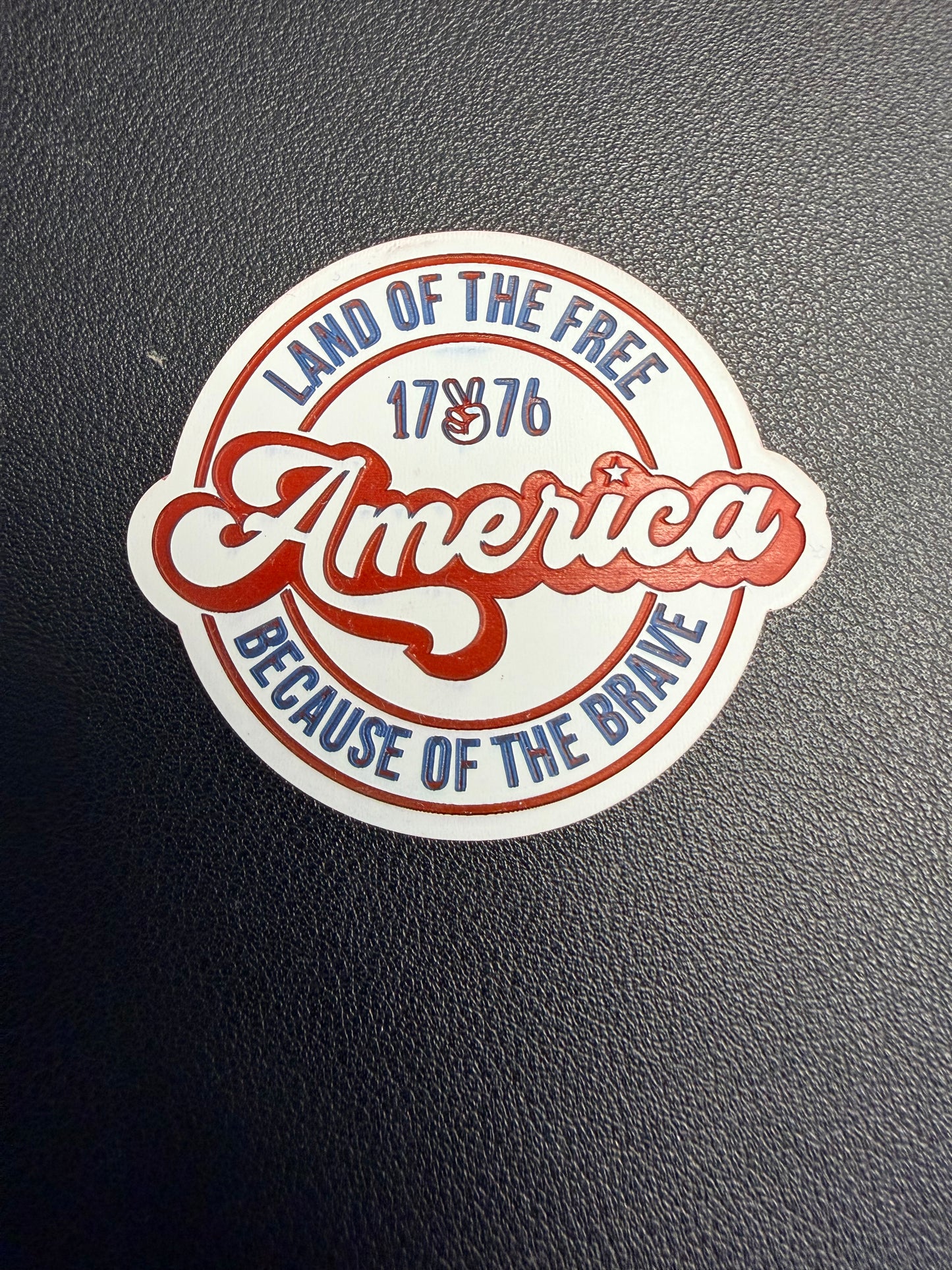 A round sticker on a textured black surface reads “America” in red script, with “Land of the Free Because of the Brave” and “1776” in blue and red—perfect inspiration for DB Custom Creations’ Custom Laser Engraved Tri Layered Acrylic Patches for Hats.
