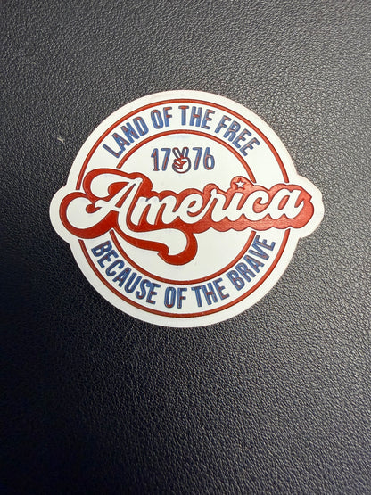 A round sticker on a textured black surface reads “America” in red script, with “Land of the Free Because of the Brave” and “1776” in blue and red—perfect inspiration for DB Custom Creations’ Custom Laser Engraved Tri Layered Acrylic Patches for Hats.