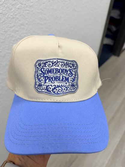 A person holds a beige and blue baseball cap featuring a DB Custom Creations Custom Laser Engraved Tri Layered Acrylic Patch on the front that says "Somebody's Problem," indoors near a white textured wall.