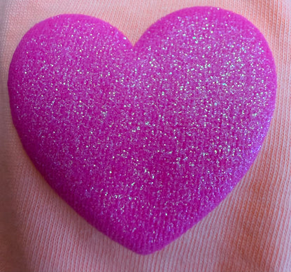A detailed close-up of a glittery pink heart, showcasing DB Custom Creations’ Custom Puff Transfers for T-Shirts and Apparel, displayed on a pale pink fabric background—perfect for adding textured designs to your clothing.