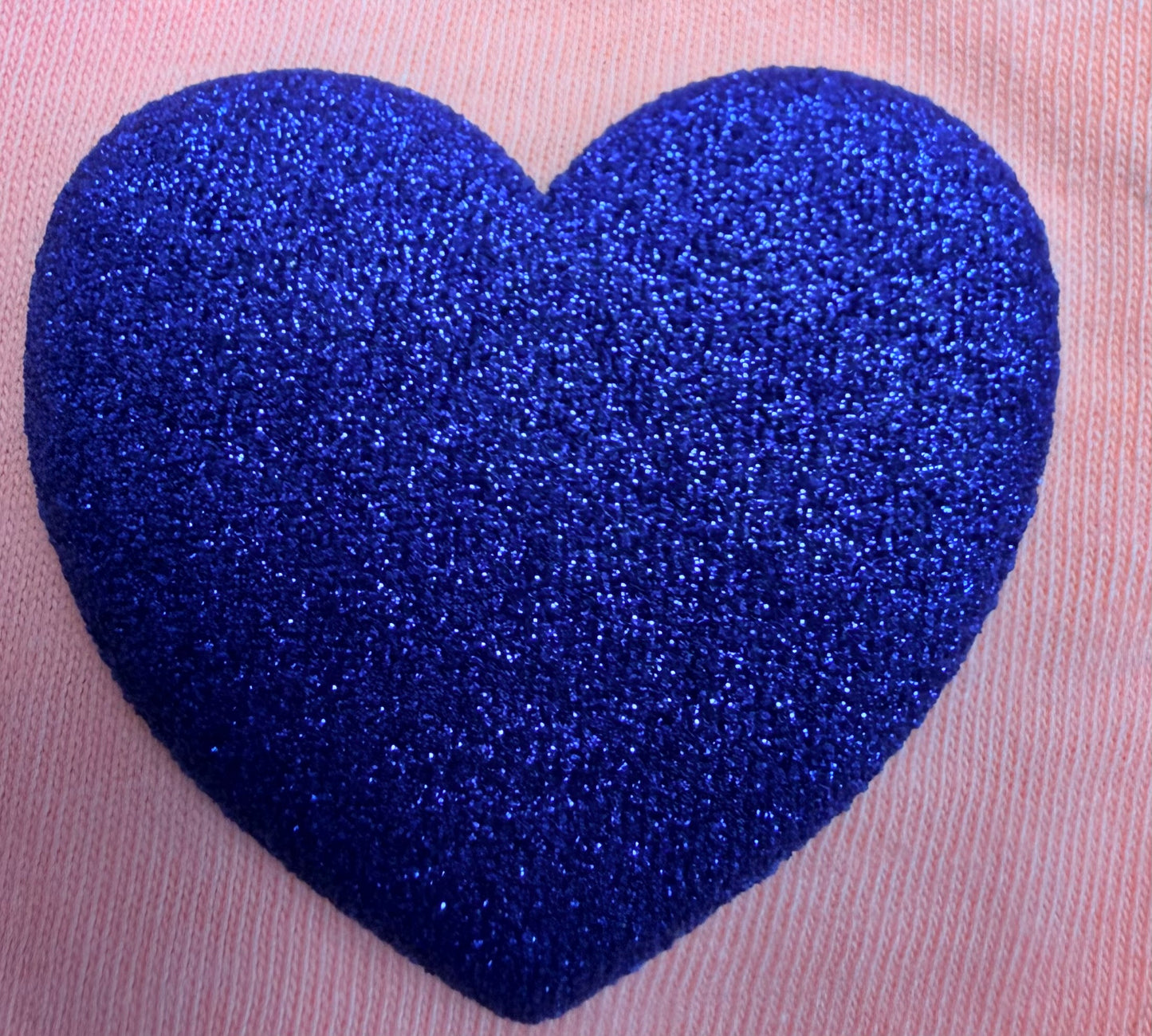 A glittery, textured blue heart stands out against a soft pink fabric background, making it an ideal design for customizing T-shirts or other apparel with DB Custom Creations’ Custom Puff Transfers for T-Shirts and Apparel.