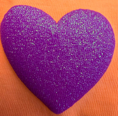 A shimmering purple heart design rests on an orange textured backdrop, making DB Custom Creations’ Custom Puff Transfers for T-Shirts and Apparel ideal for standout shirts and unique fashion statements.