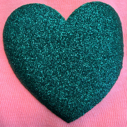 A textured, glittery dark green heart made with DB Custom Creations' Custom Puff Transfers for T-Shirts and Apparel stands out beautifully on a pink fabric background, making it an ideal accent for custom clothing or T-shirts.
