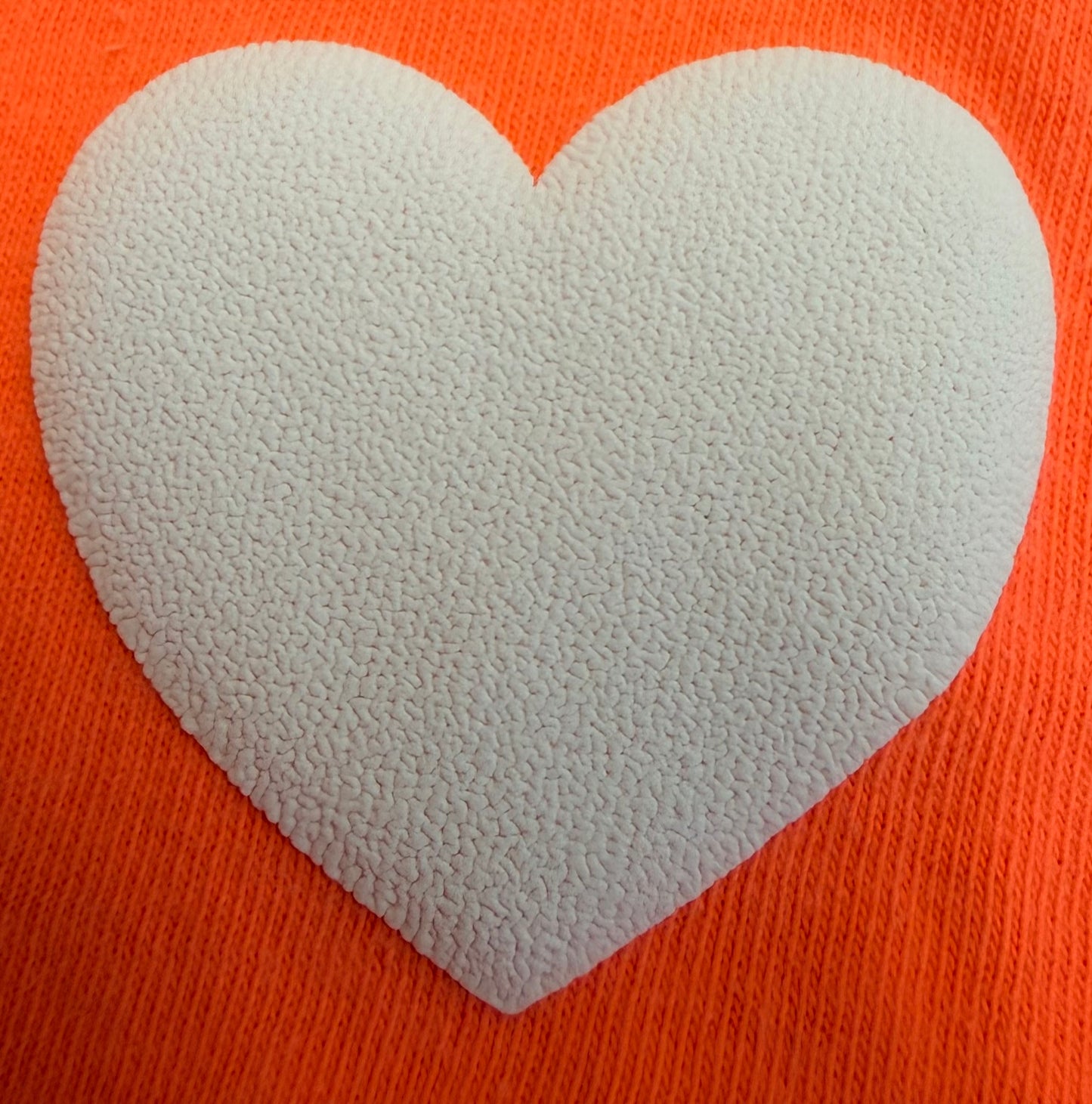 A textured white heart, crafted using DB Custom Creations' Custom Puff Transfers for T-Shirts and Apparel, pops against a vibrant orange background—ideal for giving your garments a fun and playful flair.