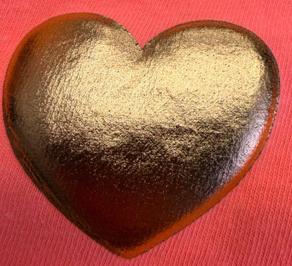 A shiny, gold-colored heart-shaped object rests on a textured pink fabric background, evoking the striking detail featured in DB Custom Creations' Custom Puff Transfers for T-Shirts and Apparel.
