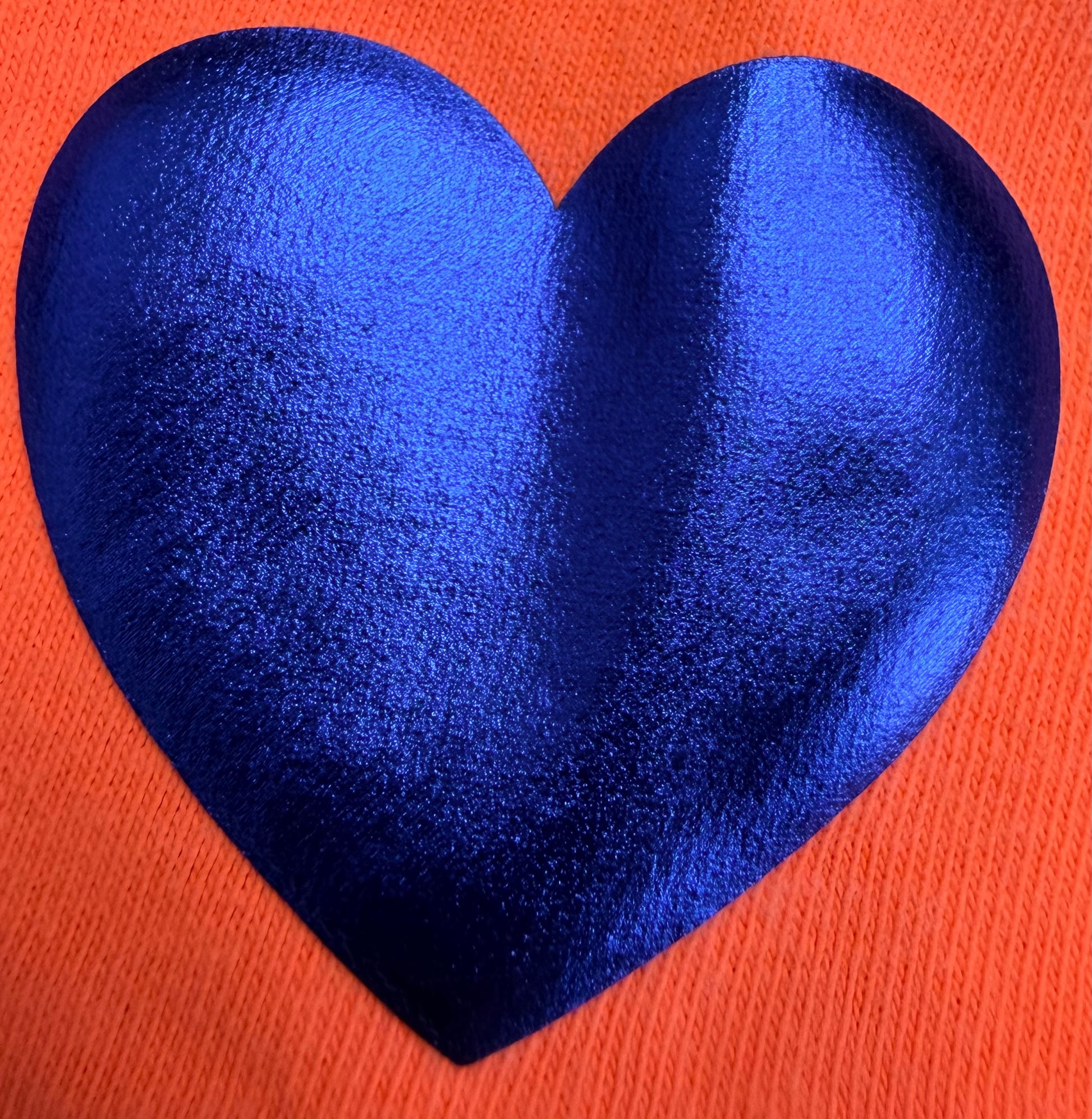 A vibrant blue heart shape, made with DB Custom Creations’ Custom Puff Transfers for T-Shirts and Apparel, stands out against a textured, bright orange fabric—ideal for creating unique and bold personalized garments.
