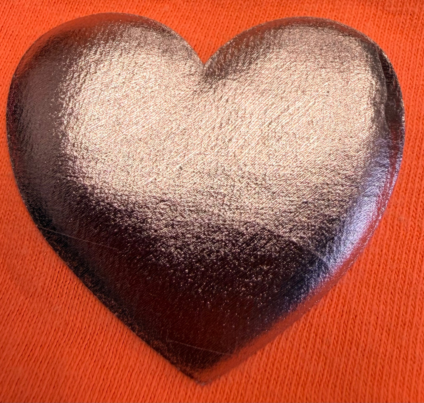 A shiny, metallic silver heart—made with DB Custom Creations' Custom Puff Transfers for T-Shirts and Apparel—stands out against a textured, bright orange fabric background, making it perfect for adding standout style to your clothing.