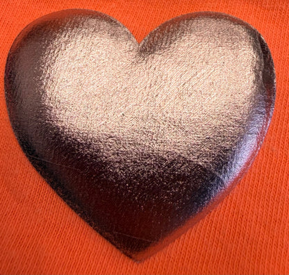 A shiny, metallic silver heart—made with DB Custom Creations' Custom Puff Transfers for T-Shirts and Apparel—stands out against a textured, bright orange fabric background, making it perfect for adding standout style to your clothing.