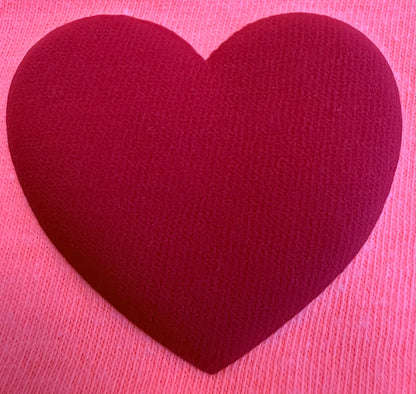 A large, textured red heart sits centered on a soft pink knitted fabric background, capturing the cozy charm featured in DB Custom Creations' Custom Puff Transfers for T-Shirts and Apparel.